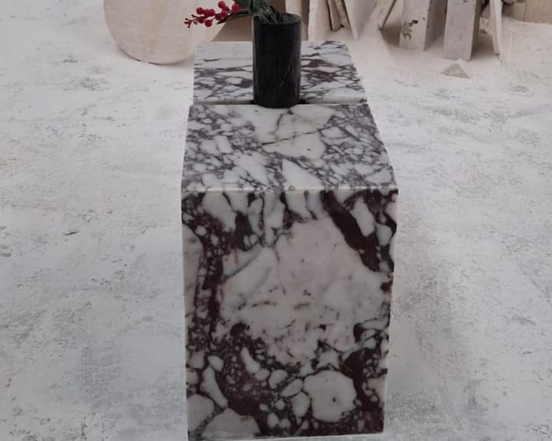 May include: A rectangular block table made of white marble with prominent dark purple and black veining. A small, cylindrical black marble vase sits on the top surface, containing a single stem with small red berries at the very top.