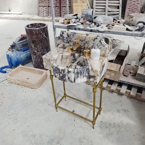 May include: A marble pedestal sink featuring a white and dark gray veined basin with a fluted exterior design. The sink is supported by a gold-toned metal frame with four legs and includes a gold faucet and a white soap dispenser on the counter.