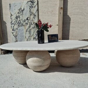 May include: A white oval marble coffee table with three round travertine bases. The table is sitting on a concrete floor in front of a wall with a marble slab.