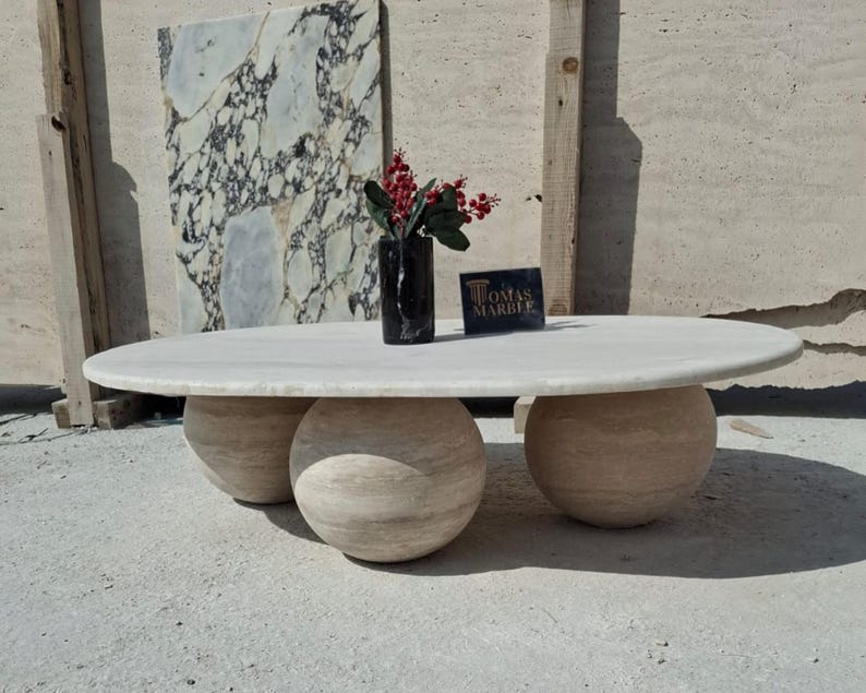 May include: A white oval marble coffee table with three round travertine bases. The table is sitting on a concrete floor in front of a wall with a marble slab.