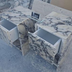 Puede incluir: Dos mesitas de noche de m&aacute;rmol con cajones abiertos. El m&aacute;rmol tiene una base blanca con vetas grises. Los cajones son blancos. Una balda de cristal con el texto "TOMAS MARBLE" est&aacute; entre las mesitas.