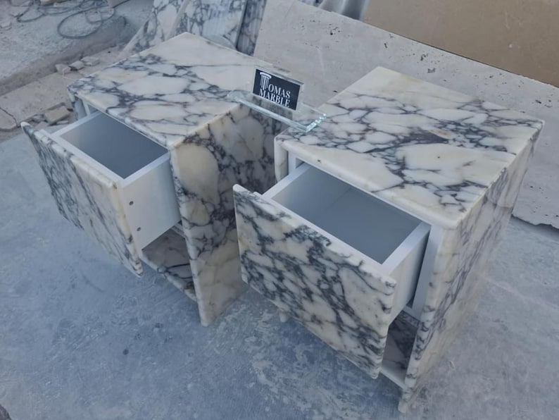 Puede incluir: Dos mesitas de noche de m&aacute;rmol con cajones abiertos. El m&aacute;rmol tiene una base blanca con vetas grises. Los cajones son blancos. Una balda de cristal con el texto "TOMAS MARBLE" est&aacute; entre las mesitas.