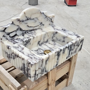 Puede incluir: Un lavabo rectangular tallado en m&aacute;rmol blanco con vetas negras prominentes. La pieza presenta un dise&ntilde;o exterior acanalado, un salpicadero alto y arqueado, y una tapa de desag&uuml;e circular de tono dorado en el fondo de la cubeta.