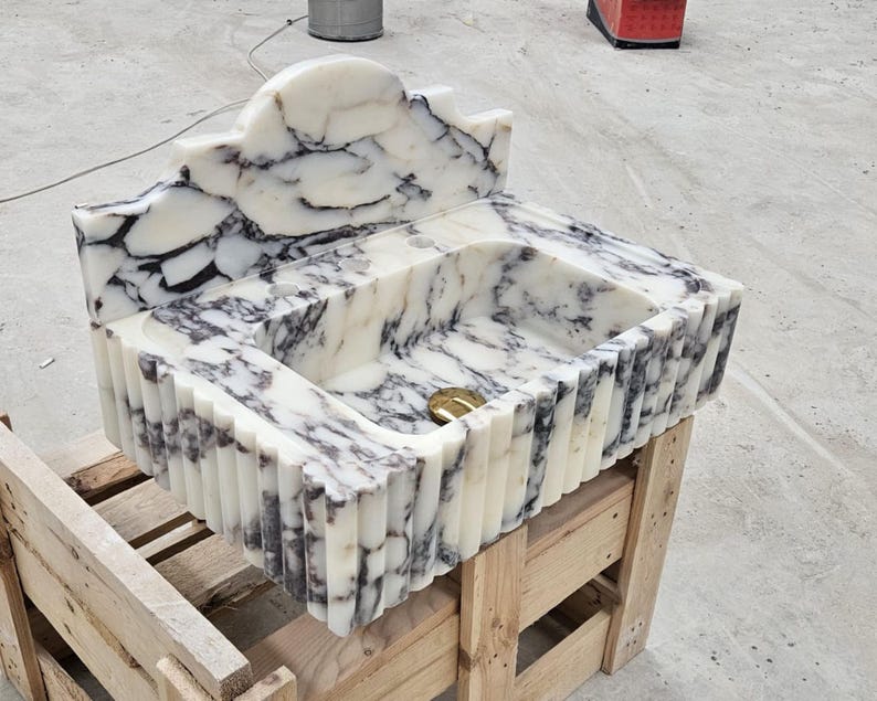 Puede incluir: Un lavabo rectangular tallado en m&aacute;rmol blanco con vetas negras prominentes. La pieza presenta un dise&ntilde;o exterior acanalado, un salpicadero alto y arqueado, y una tapa de desag&uuml;e circular de tono dorado en el fondo de la cubeta.