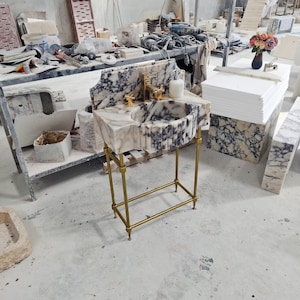 May include: A freestanding bathroom vanity featuring a white and dark purple veined marble basin with a fluted front design. The basin is supported by a slender gold-toned metal frame with four legs and a lower support bar, paired with a gold faucet.