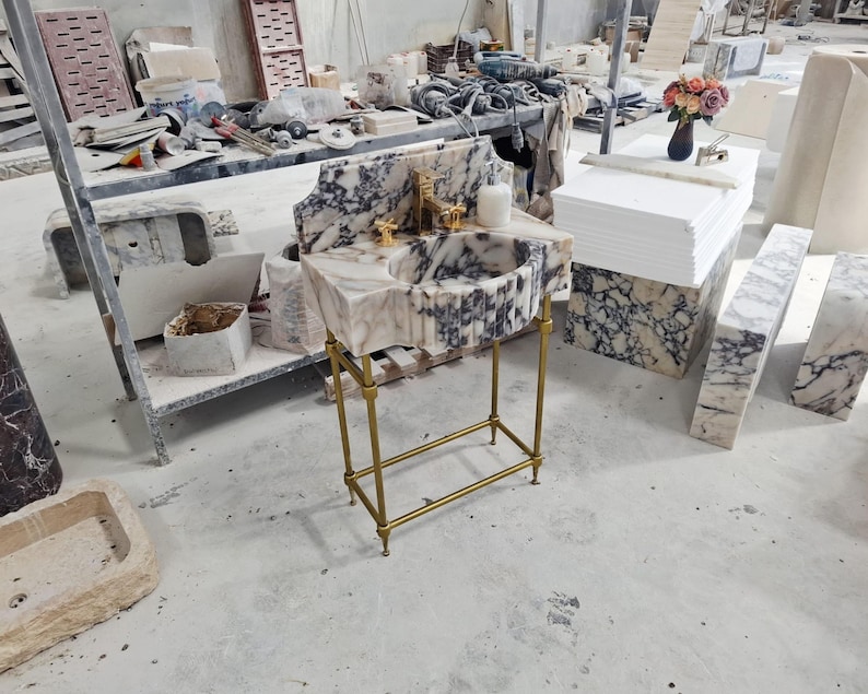 May include: A freestanding bathroom vanity featuring a white and dark purple veined marble basin with a fluted front design. The basin is supported by a slender gold-toned metal frame with four legs and a lower support bar, paired with a gold faucet.