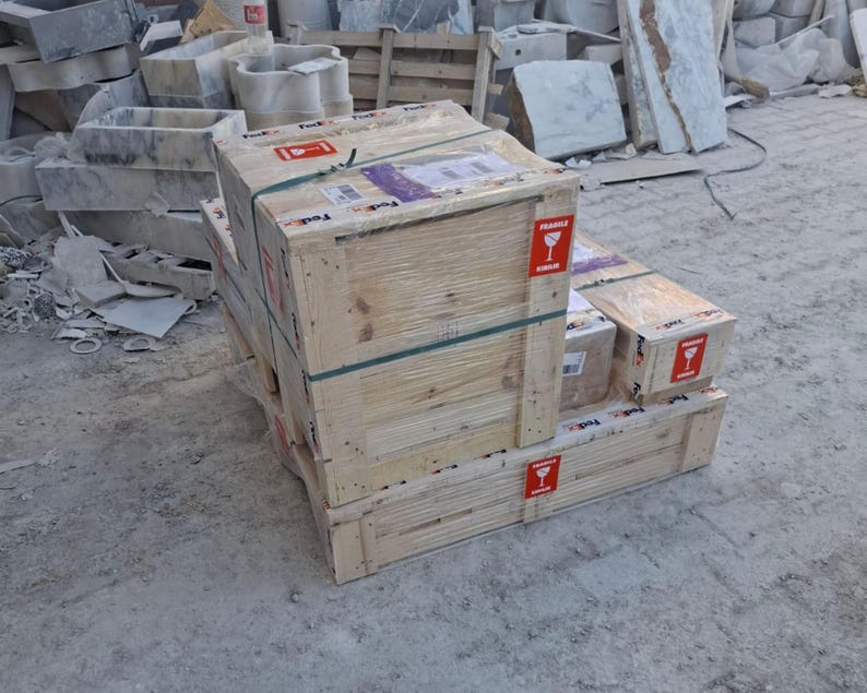 May include: A wooden shipping crate secured with plastic straps and clear shrink wrap, featuring red and white fragile labels with a wine glass icon. The crate is constructed from light-coloured pine planks and sits on a matching wooden pallet base.