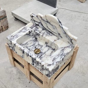 Puede incluir: Un lavabo de m&aacute;rmol blanco con vetas gris oscuro, un borde frontal festoneado y un panel trasero alto integrado. La cubeta incluye un orificio de drenaje circular central con una tapa de drenaje de metal dorado pulido.