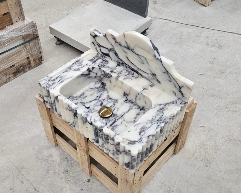 Puede incluir: Un lavabo de m&aacute;rmol blanco con vetas gris oscuro, un borde frontal festoneado y un panel trasero alto integrado. La cubeta incluye un orificio de drenaje circular central con una tapa de drenaje de metal dorado pulido.