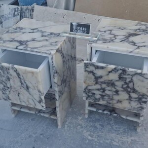 Puede incluir: Dos mesitas de noche de m&aacute;rmol con cajones abiertos. Las mesitas son blancas con vetas grises. Cada mesita tiene un caj&oacute;n y una balda. Las mesas son de m&aacute;rmol y tienen un dise&ntilde;o moderno. El texto "TOMAS MARBLE" es visible.