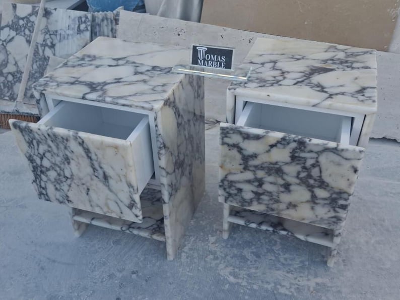 Puede incluir: Dos mesitas de noche de m&aacute;rmol con cajones abiertos. Las mesitas son blancas con vetas grises. Cada mesita tiene un caj&oacute;n y una balda. Las mesas son de m&aacute;rmol y tienen un dise&ntilde;o moderno. El texto "TOMAS MARBLE" es visible.