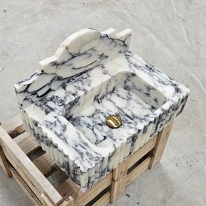 Puede incluir: Un lavabo de ba&ntilde;o de m&aacute;rmol blanco y negro con borde festoneado, un salpicadero alto y tres orificios para grifos. La cubeta incluye una tapa de desag&uuml;e circular de tono dorado pulido, mostrando vetas naturales en toda la piedra.