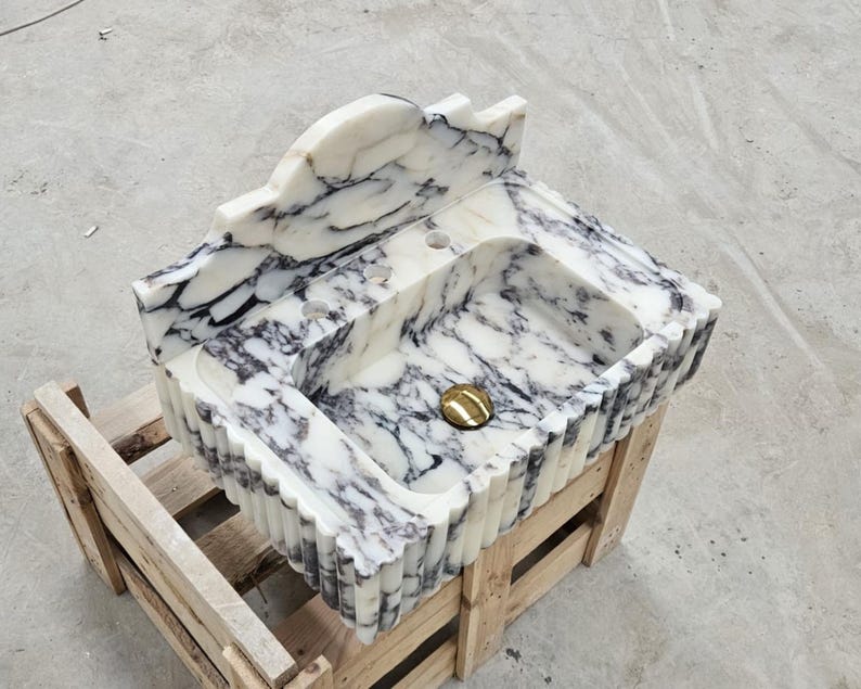 Puede incluir: Un lavabo de ba&ntilde;o de m&aacute;rmol blanco y negro con borde festoneado, un salpicadero alto y tres orificios para grifos. La cubeta incluye una tapa de desag&uuml;e circular de tono dorado pulido, mostrando vetas naturales en toda la piedra.