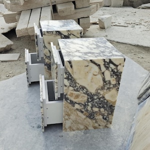May include: Two rectangular bedside tables crafted from polished stone featuring a complex, marbled pattern of cream, beige, and dark charcoal veins. Each unit includes two white drawers with a clean, modern design and a smooth, reflective surface.
