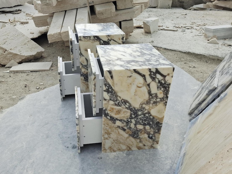 May include: Two rectangular bedside tables crafted from polished stone featuring a complex, marbled pattern of cream, beige, and dark charcoal veins. Each unit includes two white drawers with a clean, modern design and a smooth, reflective surface.
