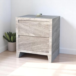 May include: A rectangular bedside table featuring two drawers with a light gray and beige marbled stone finish. The piece stands on four short legs and has a small, light-colored ceramic cup placed on its flat top surface.
