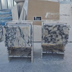 Puede incluir: Un tocador de m&aacute;rmol con dos cajones y estantes abiertos. El m&aacute;rmol tiene una base blanca con vetas grises oscuras. El tocador tiene una secci&oacute;n central espejada y est&aacute; etiquetado como "THOMAS MARBLE".