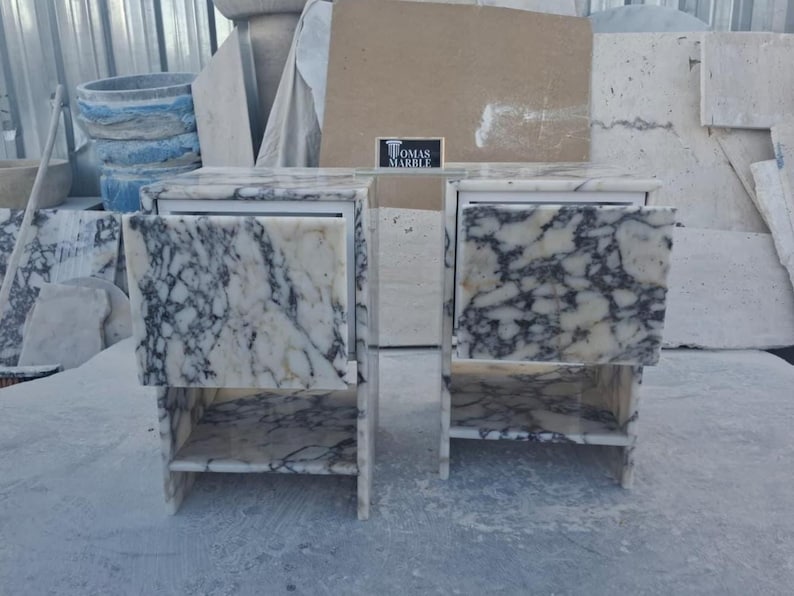 Puede incluir: Un tocador de m&aacute;rmol con dos cajones y estantes abiertos. El m&aacute;rmol tiene una base blanca con vetas grises oscuras. El tocador tiene una secci&oacute;n central espejada y est&aacute; etiquetado como "THOMAS MARBLE".