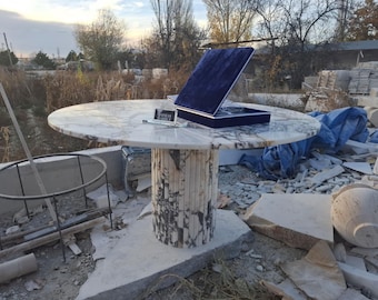 May include: A round white marble table with grey veining and a fluted cylindrical base. A small black rectangular plaque with the text "YONAS MARBLE" sits on the surface next to an open dark blue velvet presentation box containing a glass award.