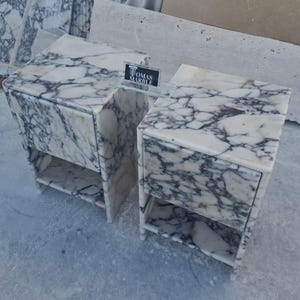 Puede incluir: Dos mesitas de noche de m&aacute;rmol con un patr&oacute;n veteado blanco y gris. Cada mesita de noche tiene un caj&oacute;n y una balda abierta. Las mesitas de noche son de m&aacute;rmol y tienen un dise&ntilde;o moderno. El letrero de la mesita de noche dice "TOMAS MARBLE".