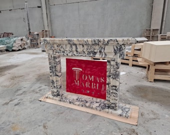 May include: A fireplace mantel crafted from white and grey veined marble, featuring a rectangular structure with a thick top shelf and vertical side pillars. A red rectangular sign is placed in the center with the white text TOMAS MARBLI.