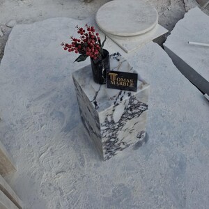 Pu&ograve; includere: Un piedistallo in marmo bianco con un piano in marmo bianco e nero. Il piedistallo &egrave; posto su una superficie bianca. Un piccolo mazzo di fiori rossi &egrave; posto sulla parte superiore del piedistallo. Un biglietto da visita nero con il testo "Thomas Marble" &egrave; posto sulla parte superiore del piedistallo.