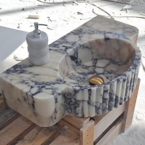 May include: A marble sink with a built-in soap dispenser. The sink has a rounded basin and a gold-colored drain. The marble has a white and gray veined pattern. The sink is on a wooden pallet.