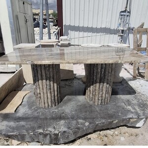 May include: A rectangular table with a polished, light brown, veined stone top. The table is supported by two cylindrical, fluted stone pedestals that match the colour and veining of the tabletop. The entire structure is made of natural stone.