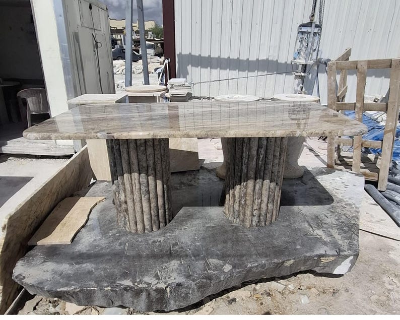 May include: A rectangular table with a polished, light brown, veined stone top. The table is supported by two cylindrical, fluted stone pedestals that match the colour and veining of the tabletop. The entire structure is made of natural stone.