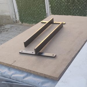 May include: Two black metal brackets are attached to a light brown piece of wood. The brackets are shaped like an L and have a flat surface on the top.