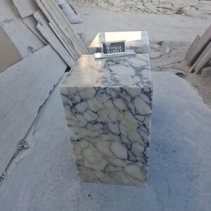 Puede incluir: Un bloque rectangular de m&aacute;rmol con un patr&oacute;n veteado blanco y gris. El bloque tiene un letrero que dice "TOMAS MARBLE". El bloque est&aacute; sobre una superficie gris.
