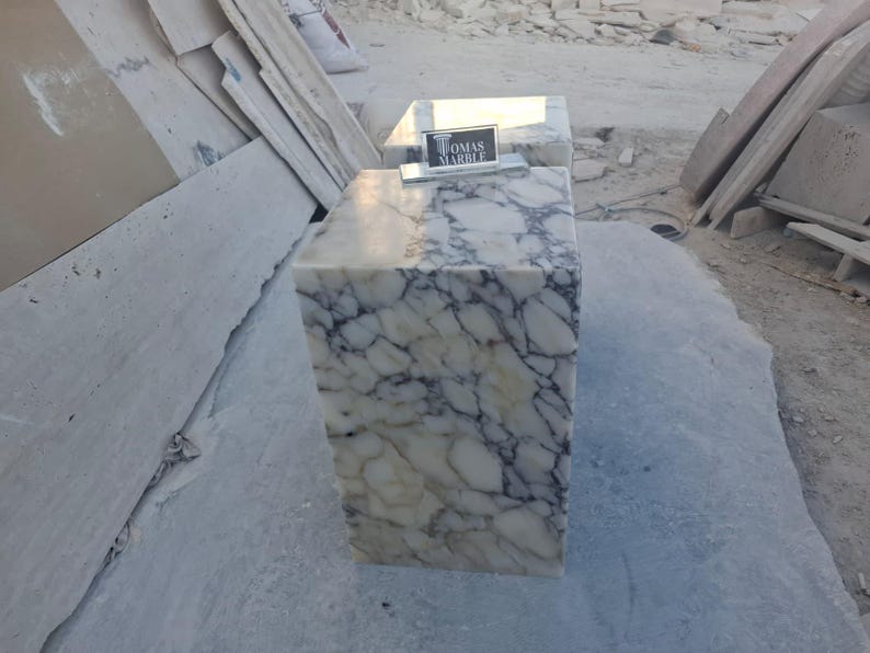 Puede incluir: Un bloque rectangular de m&aacute;rmol con un patr&oacute;n veteado blanco y gris. El bloque tiene un letrero que dice "TOMAS MARBLE". El bloque est&aacute; sobre una superficie gris.