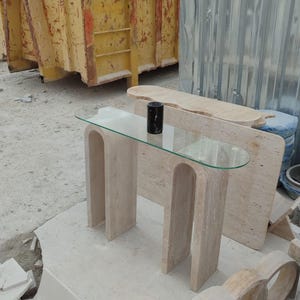 May include: A beige travertine console table with a glass top and a rounded edge. The table has two legs with a unique arch design. The table is sitting on a concrete surface.