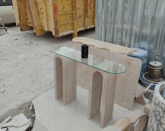 May include: A beige travertine console table with a glass top and a rounded edge. The table has two legs with a unique arch design. The table is sitting on a concrete surface.