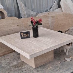 May include: A square travertine coffee table with a raised edge and a square base. The table is made of light beige travertine with a natural finish. The table is sitting on a concrete floor. The text "Tomas Marble" is visible on a small sign in front of the table.