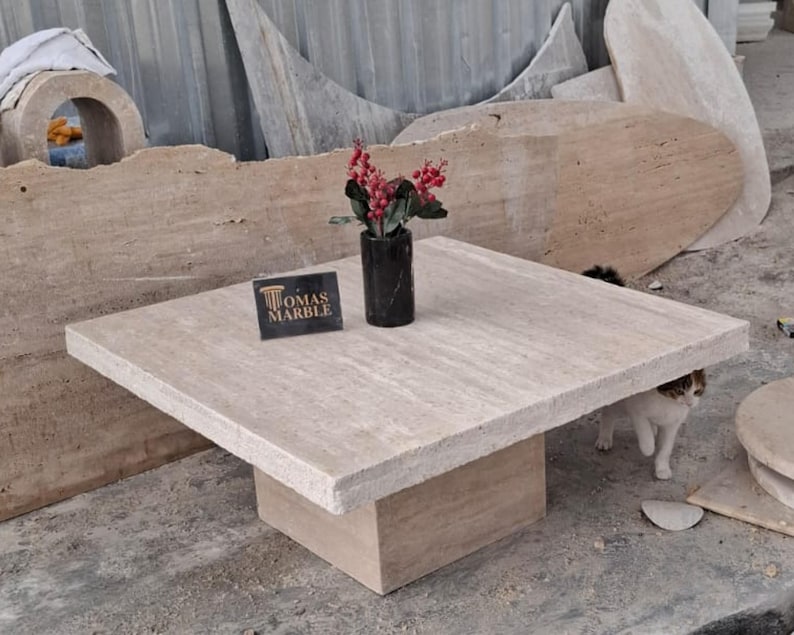 May include: A square travertine coffee table with a raised edge and a square base. The table is made of light beige travertine with a natural finish. The table is sitting on a concrete floor. The text "Tomas Marble" is visible on a small sign in front of the table.