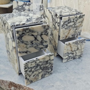 May include: Two bedside tables crafted from white and black marbled stone, each featuring two drawers with matching stone fronts. The interior drawer structures are white, and the stone surfaces display a prominent, high-contrast veined pattern.