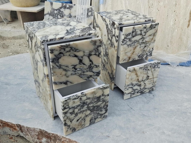 May include: Two bedside tables crafted from white and black marbled stone, each featuring two drawers with matching stone fronts. The interior drawer structures are white, and the stone surfaces display a prominent, high-contrast veined pattern.