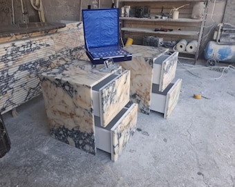 May include: Two marble-look nightstands with drawers, featuring a cream and black veined pattern. One nightstand has two drawers open, revealing white interiors. A blue case sits on top of one nightstand. The nightstands are in a workshop setting.