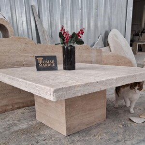 May include: A square, light beige travertine coffee table with a thick, textured top and a square base. The table is sitting on a light beige travertine floor. A white cat with brown markings is sitting under the table. A sign that reads "Tomas Marble" is sitting on the table.