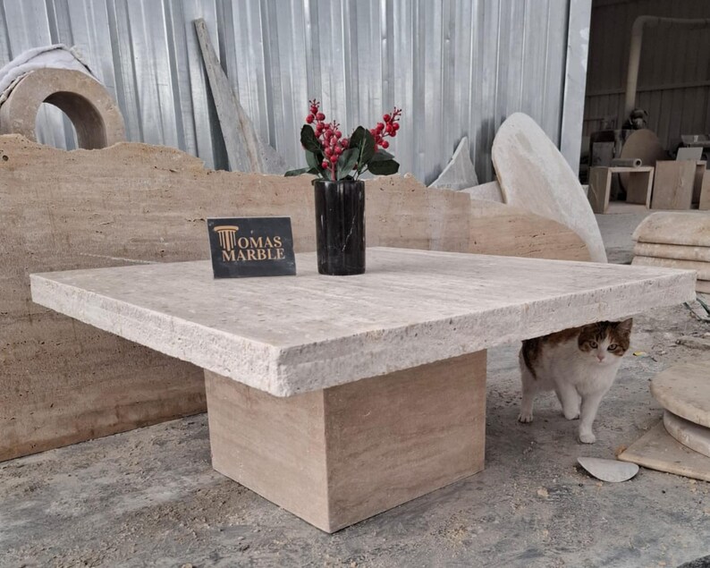 May include: A square, light beige travertine coffee table with a thick, textured top and a square base. The table is sitting on a light beige travertine floor. A white cat with brown markings is sitting under the table. A sign that reads "Tomas Marble" is sitting on the table.