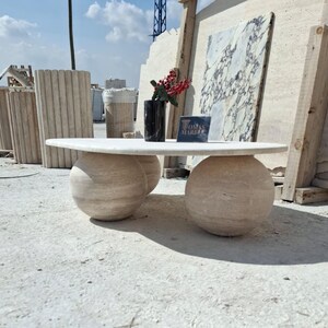 May include: A white oval-shaped coffee table with a marble top and two round marble bases. The table is sitting on a light-colored concrete floor. The text "Tomas Marble" is visible on a sign in the background.