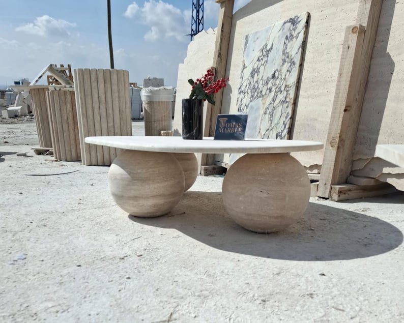 May include: A white oval-shaped coffee table with a marble top and two round marble bases. The table is sitting on a light-colored concrete floor. The text "Tomas Marble" is visible on a sign in the background.