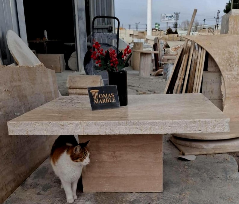 May include: A light beige travertine stone table with a square top and a rectangular base. The table is sitting on a concrete floor in a workshop setting. A cat is sitting under the table. A sign with the text "OMAS MARBLE" is on the table.