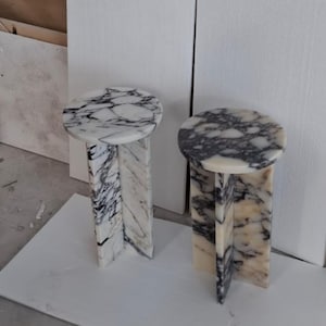 Pode incluir: Duas mesas laterais de mármore. Cada mesa tem um tampo redondo com um padrão branco e cinza. A base apresenta um design geométrico com um padrão de mármore semelhante. As mesas estão numa sala com paredes brancas e chão branco.