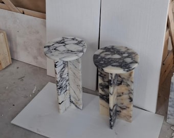 May include: Two marble side tables. Each table has a round top with a white and gray veined pattern. The base is a geometric design with a similar marble pattern. The tables are in a room with white walls and a white floor.