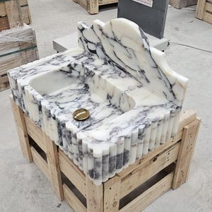Puede incluir: Un lavabo de m&aacute;rmol blanco y negro con un dise&ntilde;o exterior estriado, un salpicadero alto con borde superior curvo y una tapa de desag&uuml;e circular de tono dorado. La cubeta tiene tres orificios pretaladrados para instalar el grifo.