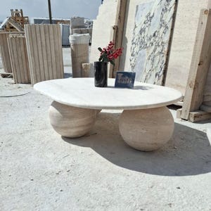 May include: A white marble coffee table with a round top and two round, ball-shaped legs. The table is sitting on a concrete surface. The text "Tomas Marble" is visible on a small sign on the table.