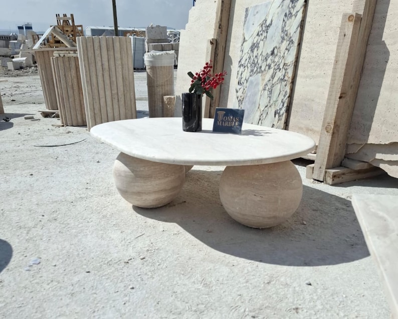 May include: A white marble coffee table with a round top and two round, ball-shaped legs. The table is sitting on a concrete surface. The text "Tomas Marble" is visible on a small sign on the table.