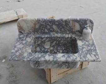 Op de afbeelding: Een marmeren wastafel met een rechthoekige wasbak en een backsplash. De wastafel heeft een grijs en wit patroon, met een kleine zeepdispenser aan de rechterkant. De wastafel staat op een houten krat.